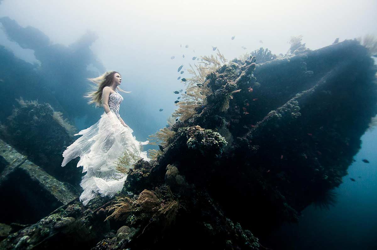 Photograph of a woman in a white dress walking the bow of a sunken ship underwater