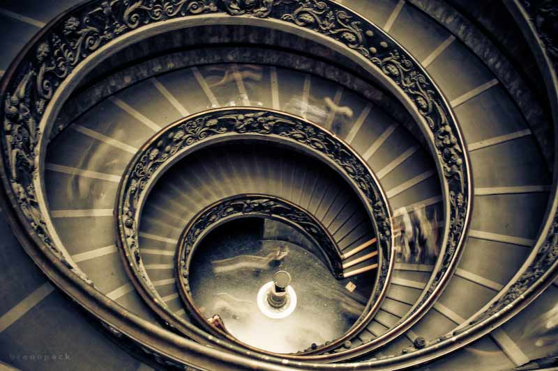 Timelapse photograph of an ornate spiral staircase in the Vatican from above, showing visitors blurring and shrinking as they descend